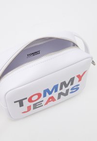Borsa cosmetica bianca con zip caratterizzata da un logo "TOMMY JEANS" ben visibile in rosso, blu e grigio. Materiale dalla texture liscia e forma rettangolare.