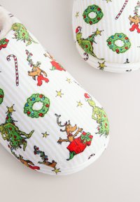 Weiße Stoffhausschuhe mit festlichem Aufdruck, der Weihnachtsbäume, Rentiere, Zuckerstangen und Kränze zeigt. Weiche Textur mit gestreiftem Muster.