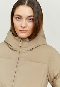 Beige padded jacket with a high collar and detachable hood, featuring a zipper closure and subtle brand logo on the chest area.