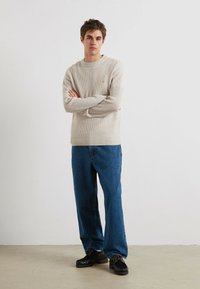 Helle beige, strukturierte Strickpullover mit Rundhalsausschnitt, kombiniert mit lässigen, locker sitzenden blauen Jeans und schwarzen Schnürschuhen.