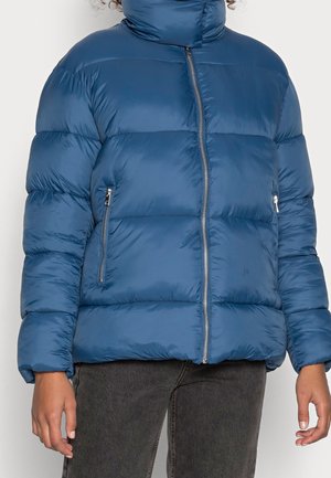 Henkilö, joka pukee sinisen toppatakin, jossa on edessä vetoketju ja kaksi vetoketjullista sivutaskua, yhdistetty tummanharmaisiin farkkuihin.