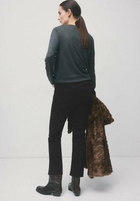 Kobieta z brązowym kucykiem, ubrana w ciemnozielony sweter, czarne spodnie i buty, trzymająca brązowy futrzany płaszcz na jednolitym jasnym tle.