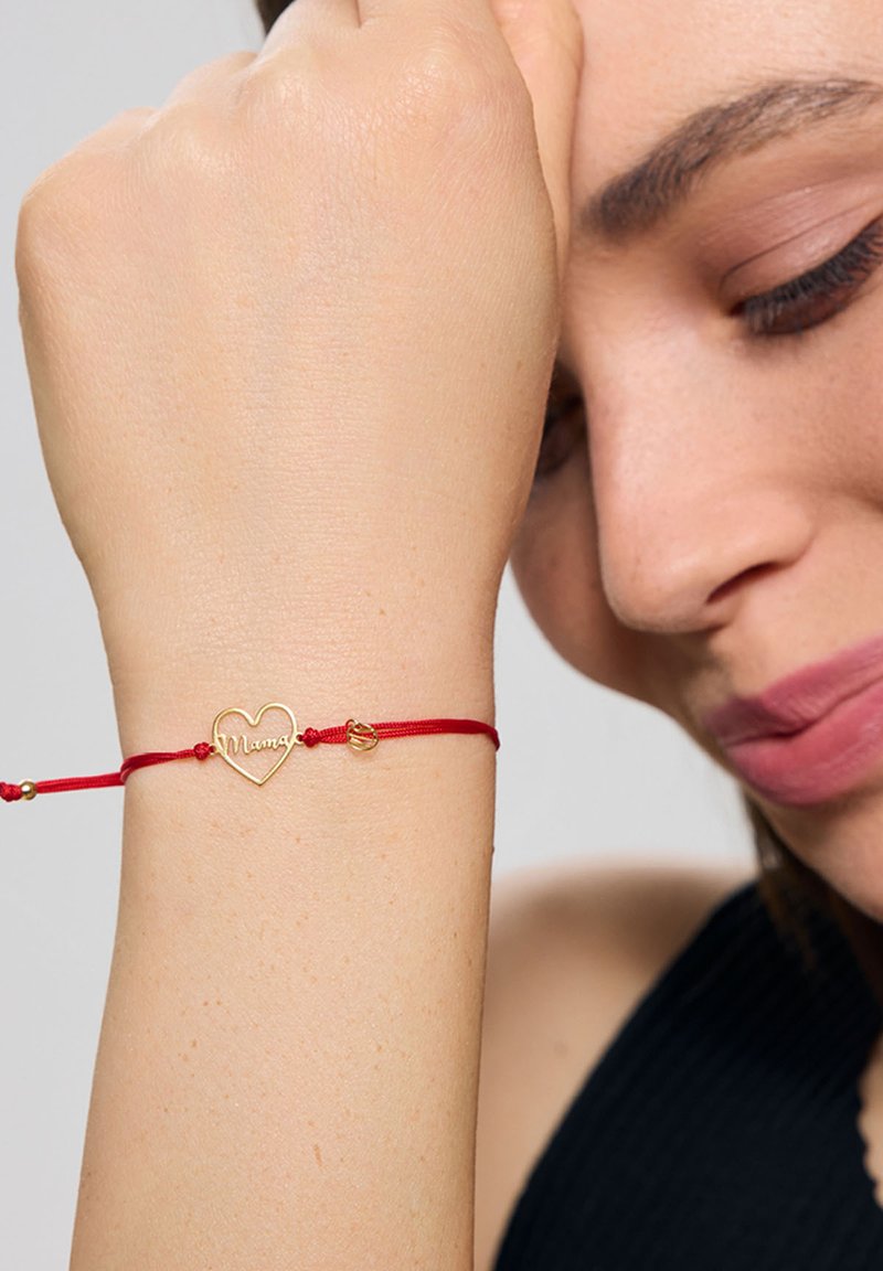 Złota bransoletka w kształcie serca z grawerem "Mama" na czerwonym sznurku, ozdobiona małymi koralikami i prostym zapięciem.