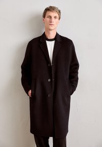 Man wearing a long black buttoned coat over a white shirt, standing with hands in pockets against a plain light wall.