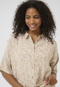 Chemise beige à manches courtes, dotée d'un motif abstrait blanc, texture lisse et col rond.