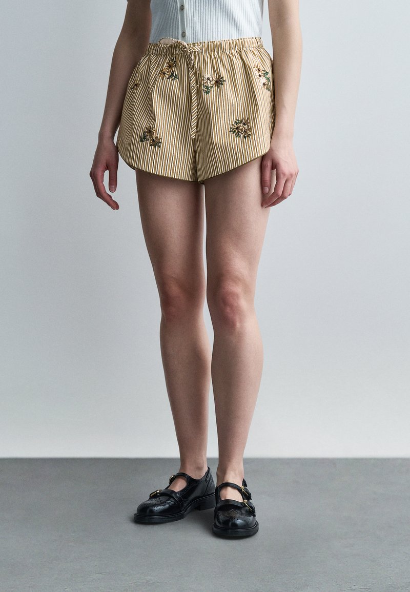 Γυναίκα που φοράει μπεζ ριγέ σορτς με κέντημα λουλουδιών, συνδυασμένα με μαύρα παπούτσια με αγκράφα, στέκεται σε γκρι πάτωμα μπροστά σε λευκό τοίχο.