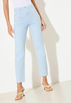 Femeie care poartă blugi albastru deschis cu talie înaltă și picior drept și sandale bej cu toc și vârf deschis, stând pe un fundal alb simplu.