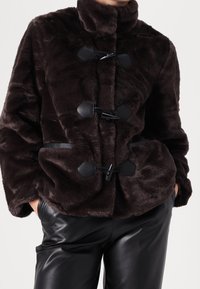 Brązowa kurtka ze sztucznego futra z wysokim kołnierzem, zapinana na troczki z czarnymi akcentami. Posiada miękką fakturę i luźny krój.