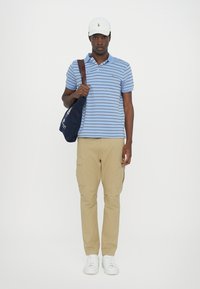 Ανοιχτό μπλε πουκαμίσα με ρίγες, καφέ cargo παντελόνι, λευκά αθλητικά παπούτσια και λευκό καπέλο. Κουβαλάει μια σκούρα μπλε τσάντα ώμου με λογότυπο.