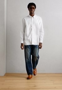 Cămașă albă cu dungi, mânecă lungă, blugi bleumarin, pantofi din piele maro cu accente metalice și ochelari de soare. Design simplu și modern.