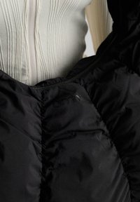 Čierna nadýchaná vesta s prešívanou textúrou, s uzáverom na zips a jemným brandingom na látke. Nosená cez svetlý, rebrovaný top.