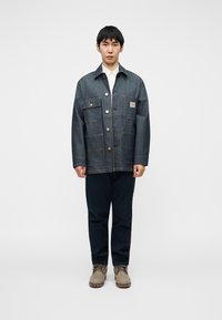 Τζιν μπουφάν με τέσσερις εμπρόσθιες τσέπες και κουμπώματα με κουμπιά, σε συνδυασμό με σκούρο μπλε παντελόνι και ανοιχτό καφέ παπούτσια. Ομαλή υφή και δομημένο σχήμα.