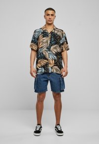 Kortærmet sort skjorte med farverigt bladprint; parret med knælange denim cargo shorts og sorte sneakers, stående mod en lys baggrund.