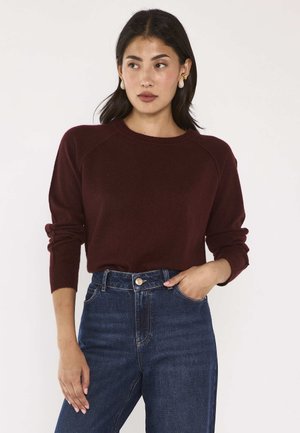Frau mit dunklen Haaren trägt einen burgunderfarbenen Pullover mit langen Ärmeln und hoch taillierte blaue Jeans, steht mit einer Hand in der Tasche vor weißem Hintergrund.