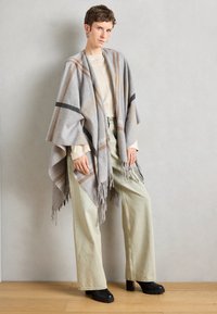 Poncho grigio in lana con frange, caratterizzato da un motivo a scacchi in beige e nero, indossato sopra una maglia beige chiaro e abbinato a pantaloni verdi larghi.