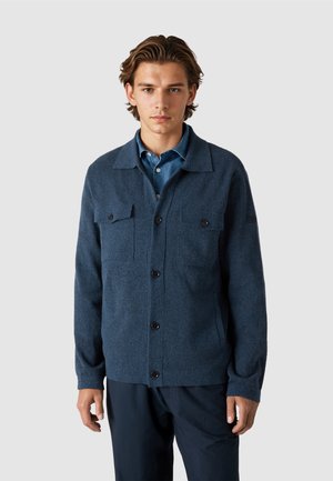 Joven vestido con una chaqueta de lana azul oscuro abotonada sobre una camisa azul con cuello, de pie frente a un fondo liso y claro.