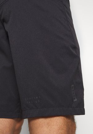 Schwarze Shorts am Oberschenkel einer Person mit blassem Text "Surfing Trails" und kleinem vertikalem Logo in der Nähe des Saums.