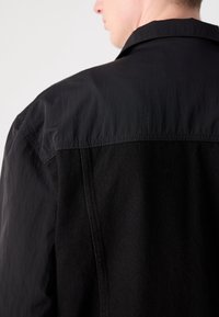 Personne portant une veste noire avec un détail de couture visible à l'arrière et une section supérieure en tissu légèrement texturé.