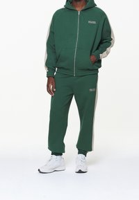 Felpa verde con zip e strisce grigie ai lati, abbinata a pantaloni della tuta verdi coordinati. Entrambi presentano una vestibilità comoda e polsini a costine. Sneakers bianche.