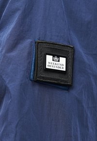 Σκούρο μπλε ύφασμα με μαύρο υφασμένο τετράγωνο με λευκή ετικέτα που γράφει "WEEKEND OFFENDER" κάτω από μικρό γραφικό κορώνας.