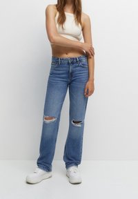 Jeans de mezclilla de un azul claro desgastados, con grandes desgarros en las rodillas, diseño de pierna recta, lavado medio, combinados con zapatillas blancas y una camiseta ajustada sin mangas.