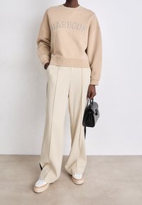 Beige sweatshirt with "BARBOUR" logo, cream wide-leg trousers, white and beige sneakers, and a small black handbag. Smooth textures throughout.
