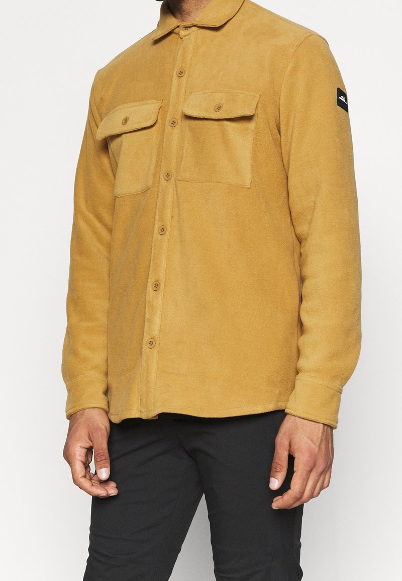 Homme portant une chemise polaire moutarde boutonnée avec deux poches poitrine et un pantalon noir, debout devant un fond uni.