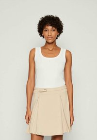 Donna con capelli ricci che indossa un top senza maniche bianco e una gonna plissettata beige con cintura, in piedi contro uno sfondo chiaro e semplice.
