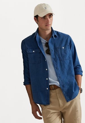 Blauwe denimoverhemd met twee borstzakken, knoopsluiting, opgerolde mouwen en een lichtblauwe T-shirt eronder, gecombineerd met beige broek.