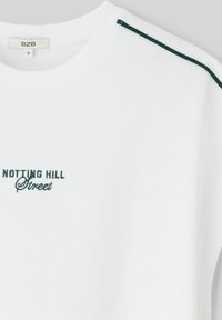 T-shirt blanche avec le texte brodé en vert "NOTTING HILL Street" sur le devant, présentant des rayures contrastantes vert foncé sur les épaules.