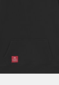 Felpa nera con cappuccio realizzata in tessuto morbido, con tasca frontale e un'etichetta del marchio rossa con testo bianco cucito sul lato inferiore.