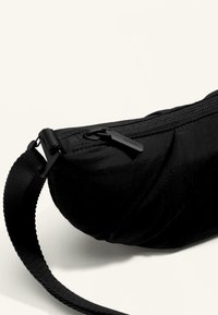 Czarna nylonowa torba w kształcie łuku, z zamkiem błyskawicznym i regulowanym paskiem. Charakteryzuje się gładką fakturą i minimalistycznym designem.