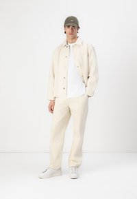 Beige knappjakke over en hvit T-skjorte, sammen med beige bukser og hvite sneakers. Laget av bomull, avslappet passform, minimalistisk design.