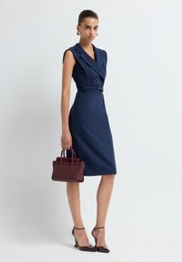 Femme dans une robe sans manches bleue marine ceinturée, tenant un sac à main bordeaux, portant des sandales à bride au niveau de la cheville bordeaux assorties et des boucles d'oreilles en or.