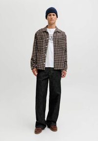 Camisa de cuadros marrón y blanca de botones sobre una camiseta gráfica blanca, combinada con unos jeans oscuros y sueltos, y unos zapatos marrones, rematada con un gorro azul.