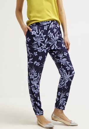 Vrouw draagt marineblauwe broek met lichtpaarse bloemenprint, gele mouwloze top en beige ballerina's met strass-steentjes.