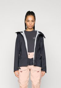 Crna vodootporna jakna sa sivoj unutrašnjosti, zatvorenim džepovima i podesivom kapuljačom, uparena sa svijetloružičastim cargo hlačama koje imaju više džepova.