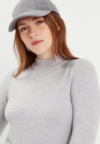 Un pull gris clair avec un col haut, une texture côtelée et trois boutons métalliques sur une épaule. Assorti à une casquette grise.