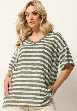 Femme blonde portant un haut ample à manches courtes avec des rayures horizontales vert olive et blanches, col en V, et un pantalon beige, sur un fond uni.