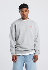 Gray sweatshirt with long sleeves, round neckline, and a small red logo on the chest. Worn over a white t-shirt, paired with light blue jeans.