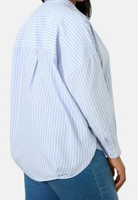 Donna che indossa una camicia a maniche lunghe a righe verticali blu chiaro e bianche e jeans blu, mostrata da dietro e di lato.