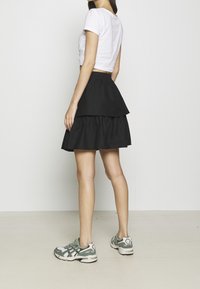 Kvinna som står på sidan klädd i en vit kortärmad T-shirt, svart lagerkjol och vit-gröna sneakers framför en enkel vit vägg.