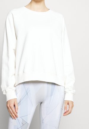 Osoba ubrana w luźną, białą bluzkę z długim rękawem oraz jasne legginsy w geometryczny wzór, stojąca na jednolitym tle.