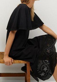 Schwarzes Kleid mit einem ausgestellten, bestickten Cape-Style-Oberteil und aufwendigen Ösenverzierungen am Saum. Hergestellt aus leichtem Stoff mit glatter Textur.