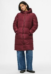 Burgundijos spalvos pūkinė striukė su užtrauktuku ir kapišonu, turinti siuvinėtą dizainą. Derinama su platėjančiomis džinsinėmis kelnėmis ir juodais auliniais batais.