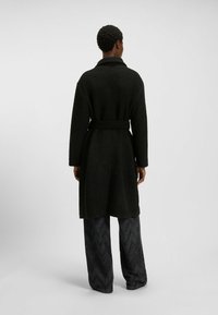 Czarny teksturowany płaszcz z paskiem w talii, z szerokim kołnierzem i długimi rękawami, połączony z wzorzystymi spodniami o szerokich nogawkach.