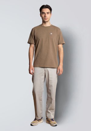 Joven de pie mirando al frente, vestido con una camiseta marrón, pantalones beige claros y zapatillas beige con cordones y suelas oscuras.