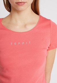 Korálově zbarvené bavlněné tričko s kulatým výstřihem a krátkými rukávy, zdobené nápisem "ESPRIT" ze stříbrných kamínků na hrudi.