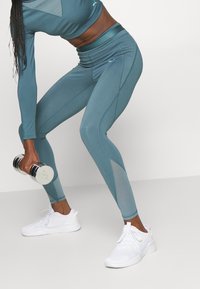 Atletický outfit obsahující modrozelený top s dlouhými rukávy a přiléhavé legíny s síťovinovými panely, doplněný bílými teniskami a držící stříbrnou činku.