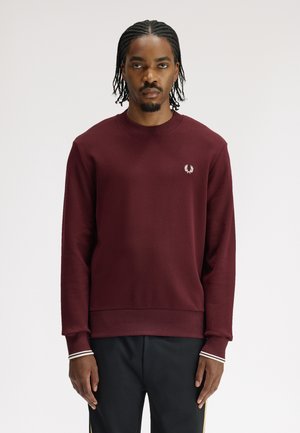 Mann mit geflochtenem Haar, der einen bordeauxfarbenen Pullover mit Rundhalsausschnitt und kleinem weißem Logo auf der linken Brust sowie schwarze Hose mit goldenen Streifen trägt.